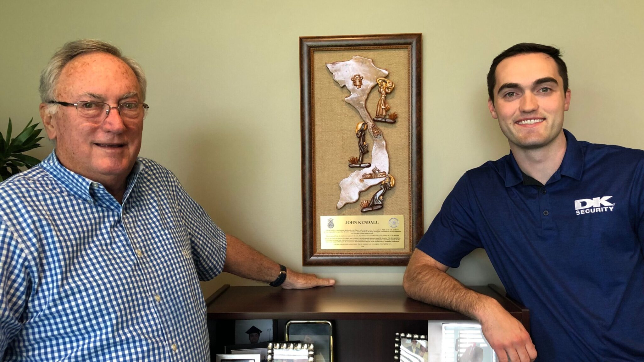 John Kendall and Johnny Kendall pose for a photo at DK Security corporate headquarters in front of a wall-mounted award recognizing John Kendall's career in the U.S. Marshals.