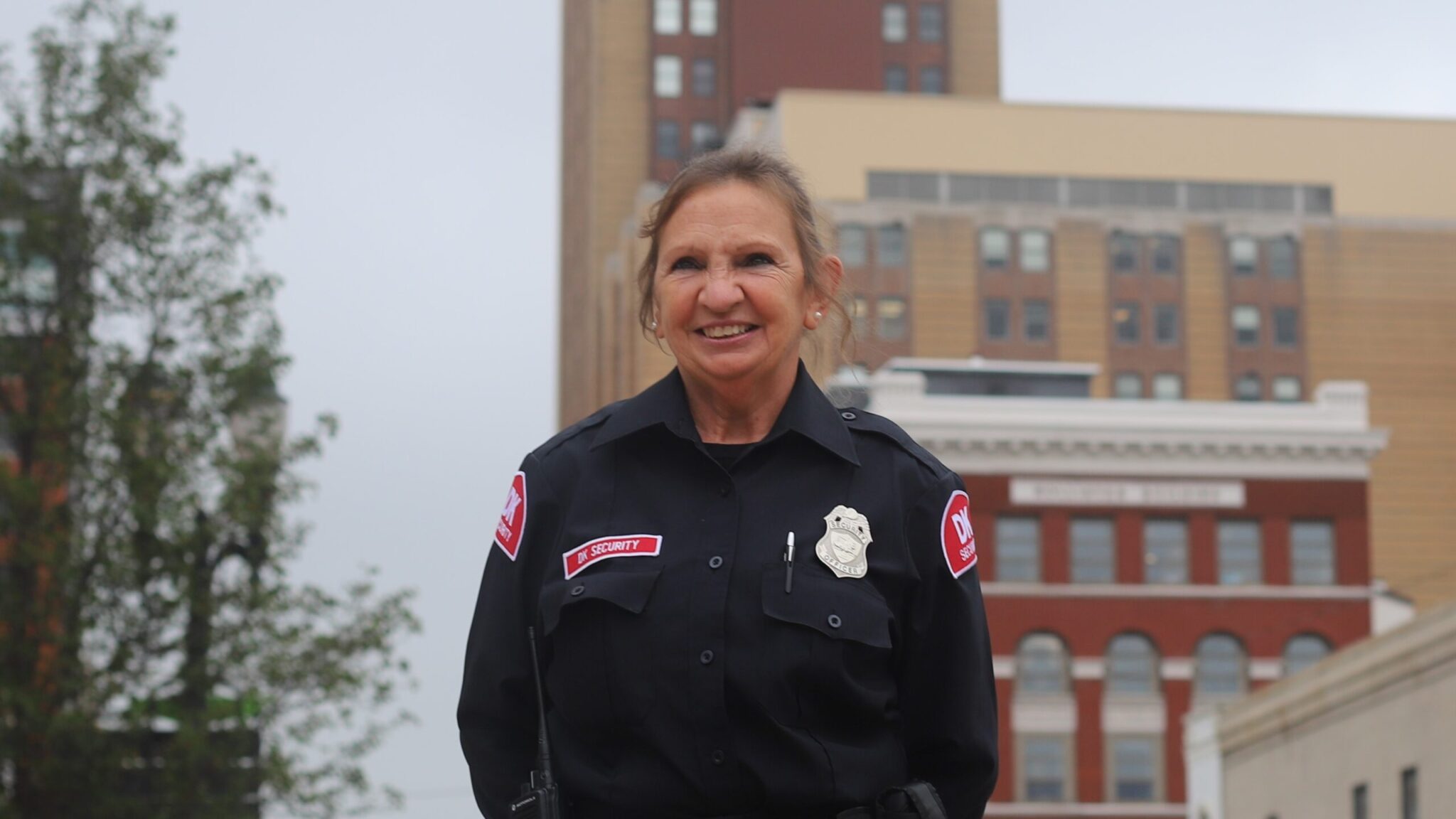 A female security guard, dressed in a DK Security navy security uniform, stands guard at a work site in downtown Lansing, Michigan. Boji Tower is partially visible in the background of the street the officer is standing on.