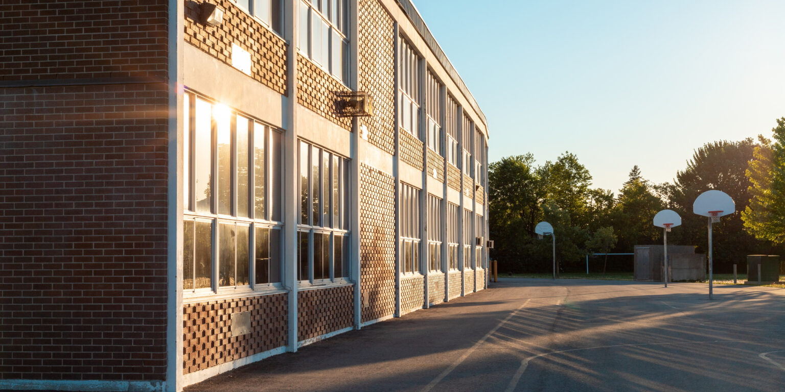 The exterior of the school building and school yard with a basketball court on a sunny evening. The sun ireflecting in the windows